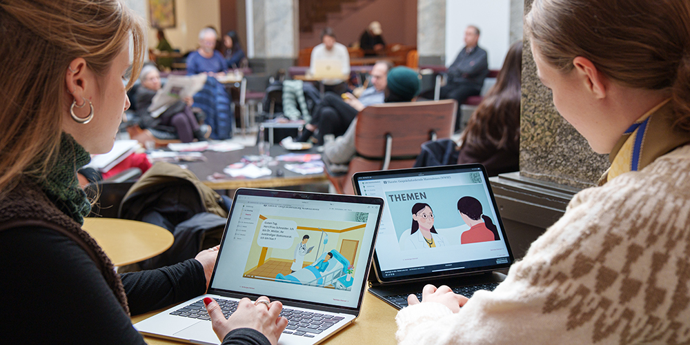 Zwei weiblich gelesene Personen sitzen mit dem Rücken zur Kamera am Tisch. Wir sehen ihre beiden Laptopbildschirme, auf denen das E-Learning abgebildet ist. Im Hintergrund sind Menschen im Kaffeehaus Unternehmen Mitte zu sehen.