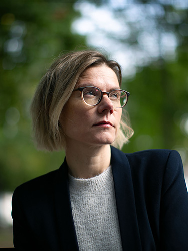 Portrait of a woman with short blonde hair and glasses, wearing a dark blazer over a light grey sweater. Portrait of a woman with short blonde hair and glasses, wearing a dark blazer over a light grey sweater.