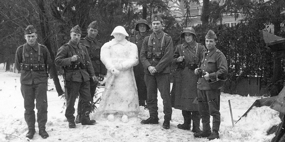 Schwarz-Weiß-Fotografie: Sieben uniformierte Soldaten stehen im Schnee um ein lebensgroßer Schneesoldat mit Helm, Gewehr und Mantel. Im Hintergrund sind Bäume und Gebäude zu sehen. Schwarz-Weiß-Fotografie: Sieben uniformierte Soldaten stehen im Schnee um ein lebensgroßer Schneesoldat mit Helm, Gewehr und Mantel. Im Hintergrund sind Bäume und Gebäude zu sehen.