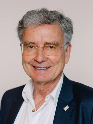 Porträt eines älteren Mannes mit grauen Haaren, Brille und dunklem Sakko über einem weißen Hemd, leicht lächelnd vor einem einfarbigen grauen Hintergrund. Porträt eines älteren Mannes mit grauen Haaren, Brille und dunklem Sakko über einem weißen Hemd, leicht lächelnd vor einem einfarbigen grauen Hintergrund.