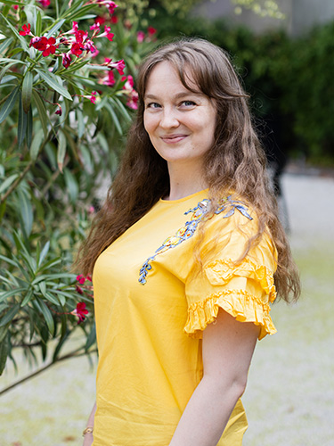 Smiling woman with long brown hair wearing a yellow top beside a flowering shrub. Smiling woman with long brown hair wearing a yellow top beside a flowering shrub.