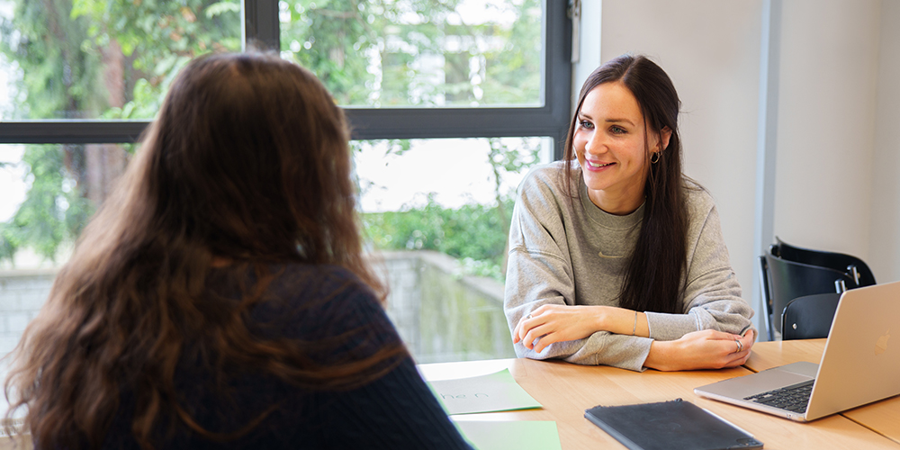 Zwei Studierende sitzen sich in einem Seminarraum gegenüber und führen ein Rollenspiel zum psychologischen Coaching durch. Auf dem Tisch liegen Unterlagen und ein Laptop.