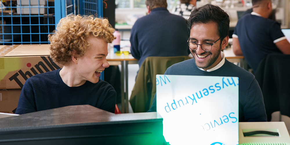 Zwei Studierende sitzen an einem Computerarbeitsplatz, lächeln und blicken auf einen Bildschirm in einer technischen Laborumgebung.