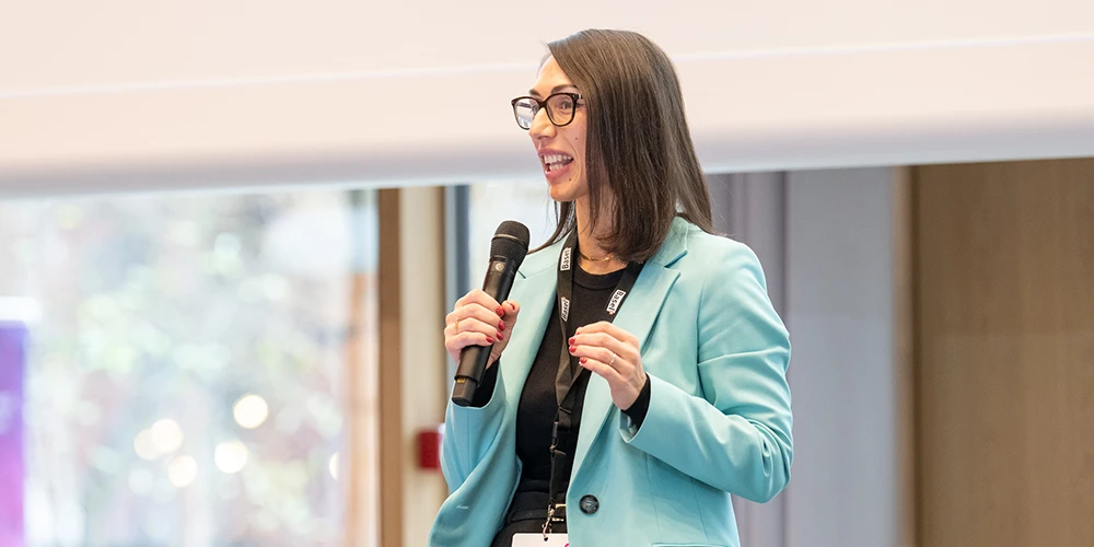 A female speaker wearing a turquoise jacket and holding a microphone addresses the audience
