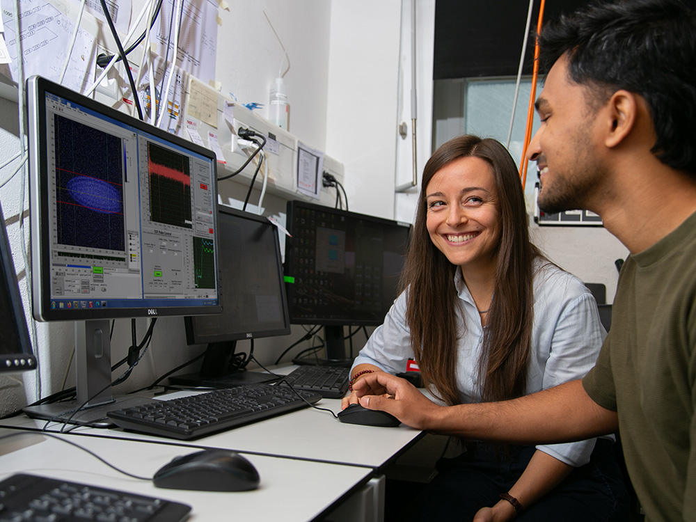 A young researcher and her doctoral student are sitting at a computer displaying a blue oval representing a Coulomb crystal. A young researcher and her doctoral student are sitting at a computer displaying a blue oval representing a Coulomb crystal.