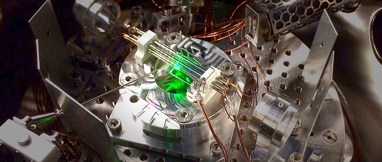 Nahaufnahme eines wissenschaftlichen Instruments mit Metallkomponenten, Drähten und einem grünen Laserstrahl in einer Vakuumkammer.